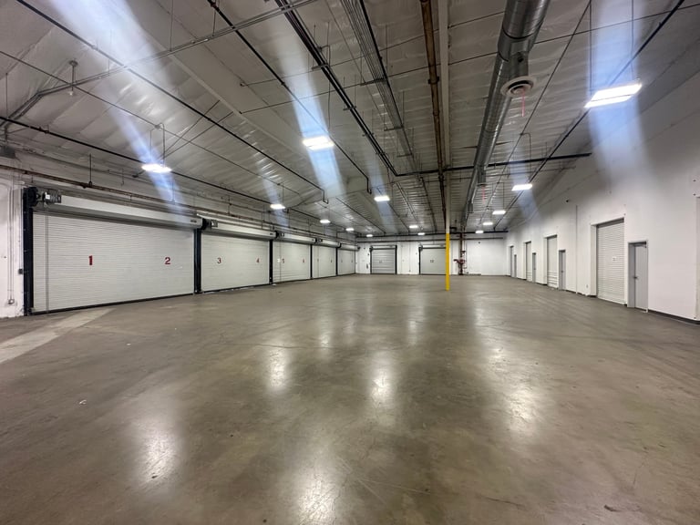 Empty industrial warehouse interior with polished concrete floor, fluorescent ceiling lights, white walls, and numbered garage doors