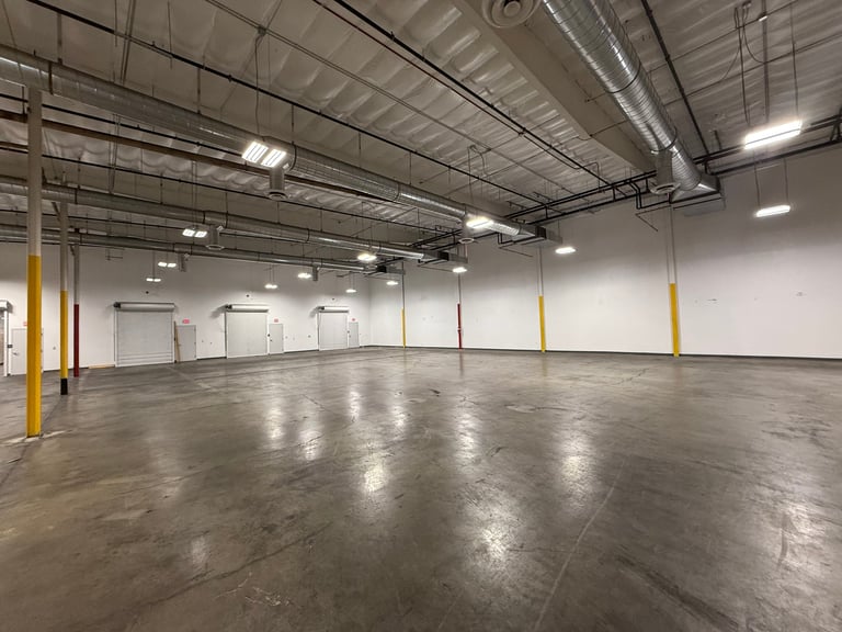 Empty warehouse interior with polished concrete floor, yellow support columns, metal ceiling with LED lights, and white walls